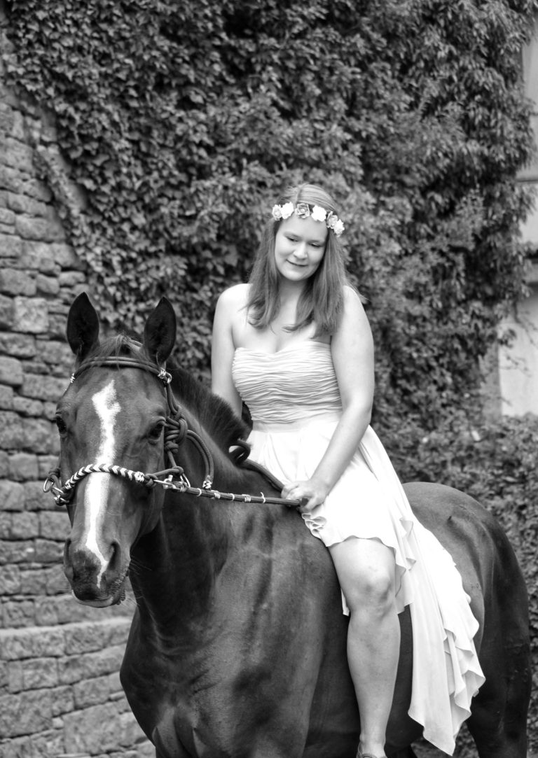 Inhaberin, wie sie auf ihrem ersten Pferd ohne Sattel sitzt. mit einem gebisslosen Zaum. 
Sie hat ein Kleid an und einen Blumenkranz im Haar